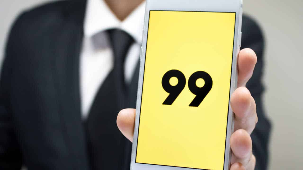 Busto de um homem de terno com celular na mão. O aparelho está apontado para a câmera com o app do 99 na tela inicial