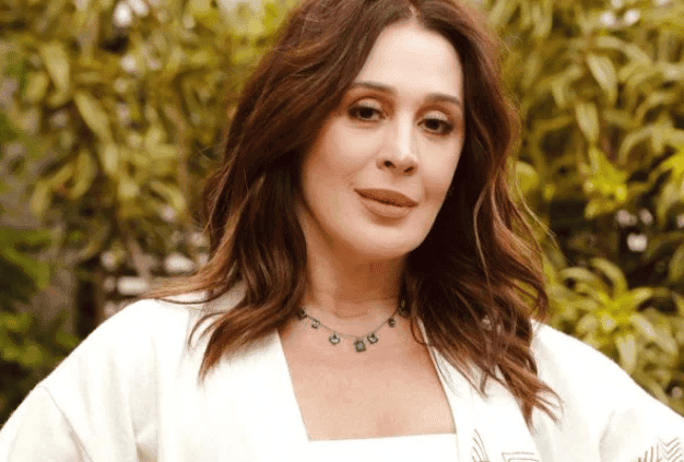 atriz Claudia Raia com blusa branca e árvores ao fundo e um colar no pescoço