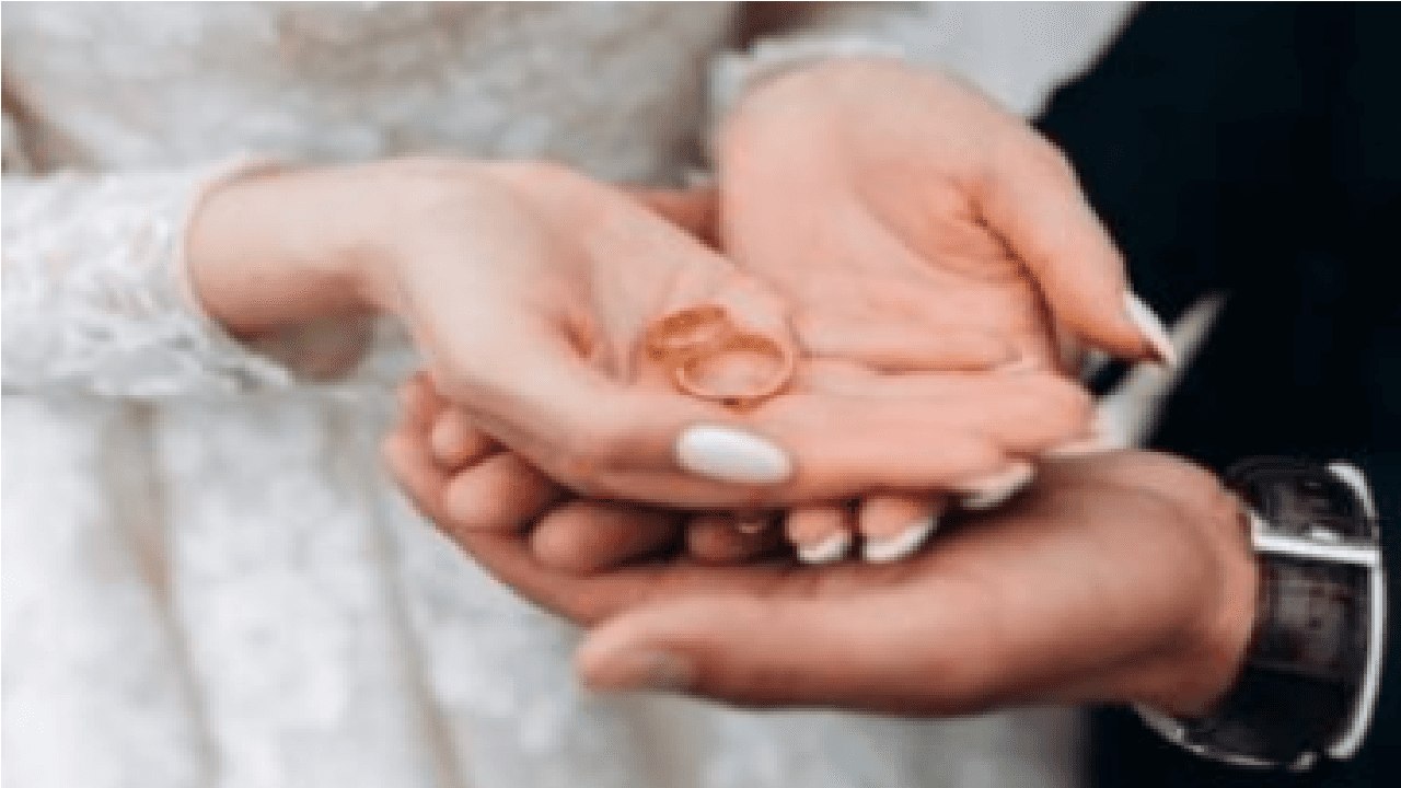 Imagem de noivo segurando as mãos da noiva, onde estão dois anéis de casamento