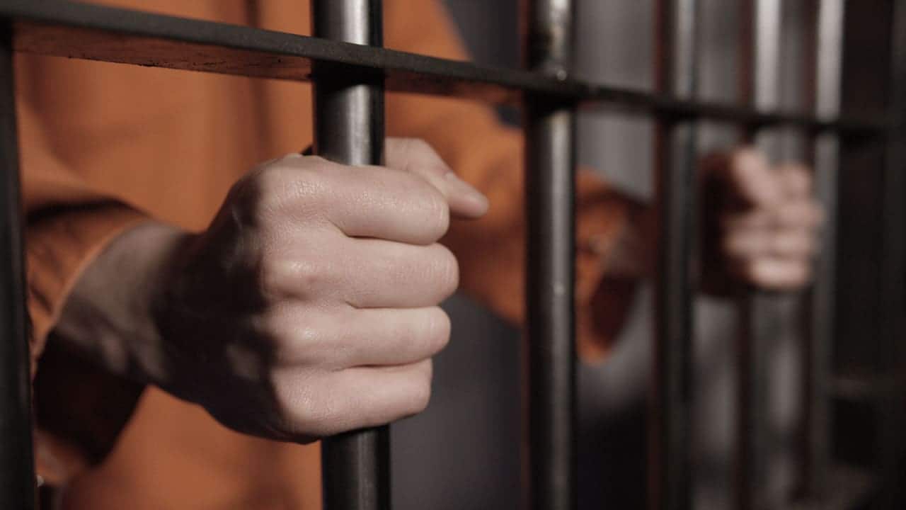 Mãos de uma pessoa segurando grades de uma cela de prisão