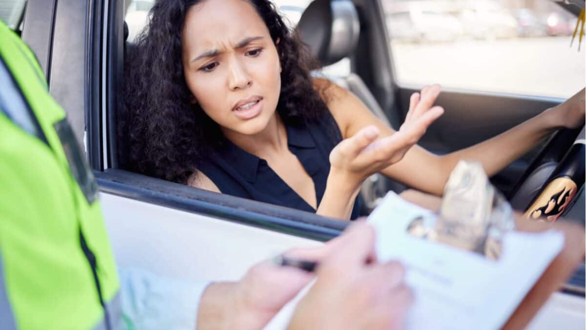 Imagem de uma mulher chateada tomando uma multa de trânsito de um agente que segura uma prancheta com um documento.