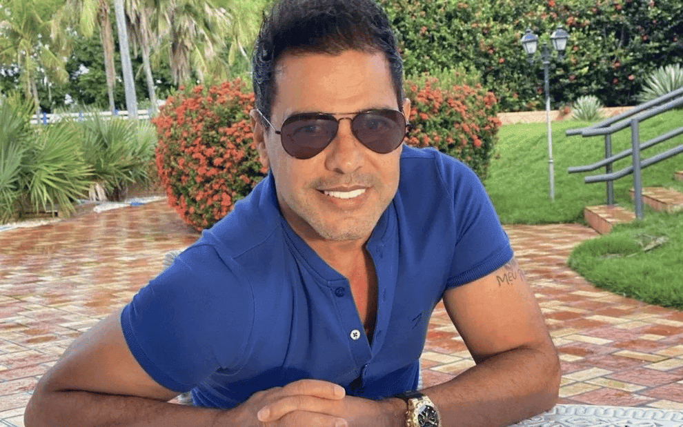 imagem do cantor Zezé di Camargo com uma camisa azul debruçado sobre um muro. Ao fundo, aparece um jardim