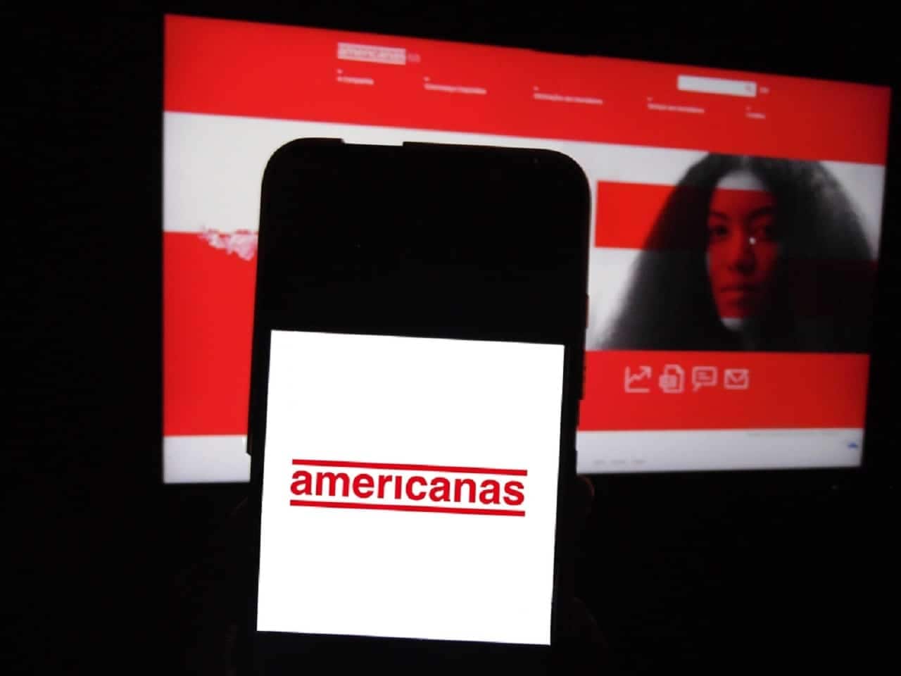Imagem de uma pessoa utilizando um celular com a tela da lojas Americanas, com computador ao fundo.