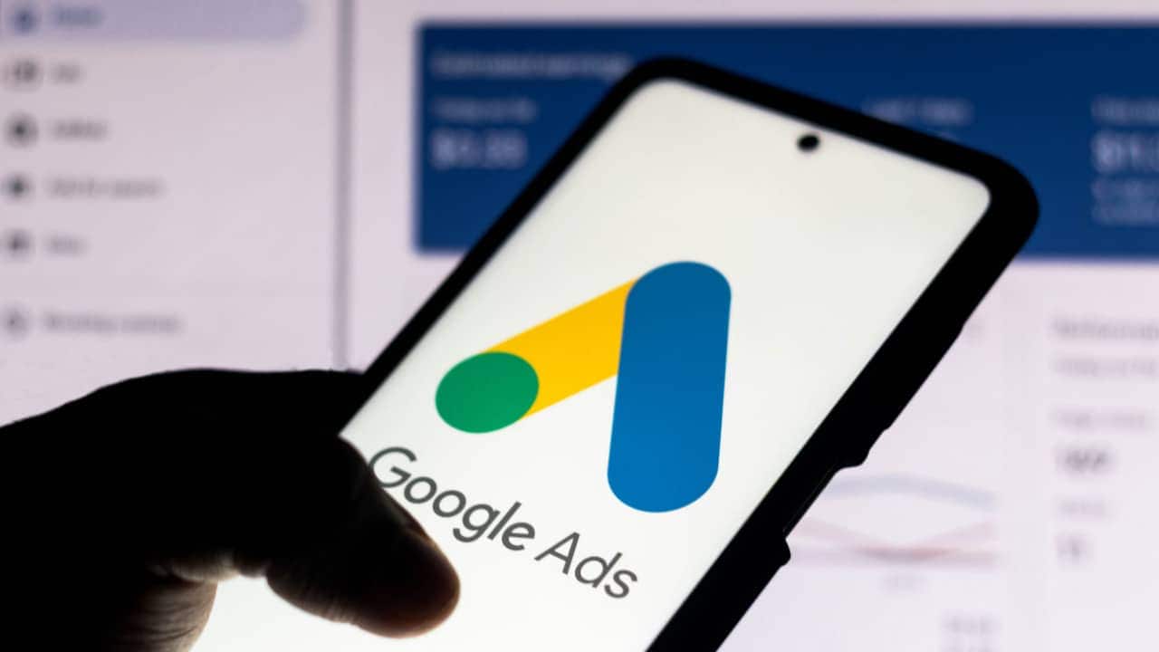 Mão segurando celular que mostra logo do Google Ads, plataforma de anúncios no Google. Ao fundo, tela com o site da ferramenta aparece desfocada.