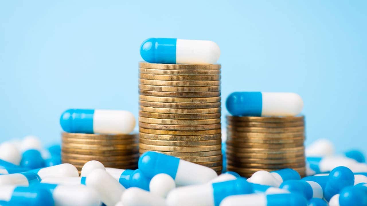 Três montes de moedas, cada uma com uma cápsula azul e branca no topo. À volta dos montes, várias cápsulas do mesmo medicamento.