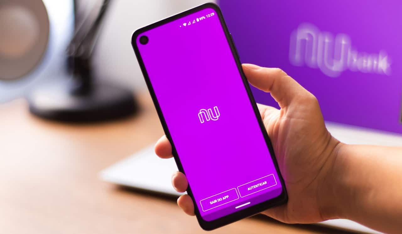 Mão segurando celular que mostra tela inicial do aplicativo do Nubank. Ao fundo, tela de computador desfocada mostra logo do banco.