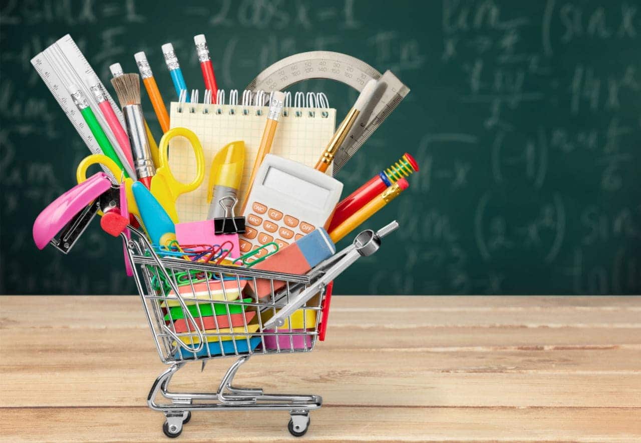Miniatura de carrinho de compras sobre uma mesa com vários materiais escolares dentro. Ao fundo, um quadro de sala de aula com anotações em giz branco