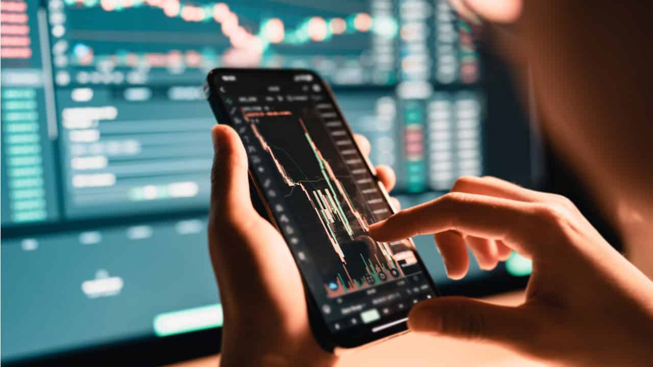 Pessoa segurando celular com bolsa de valores e tela ao fundo com ações