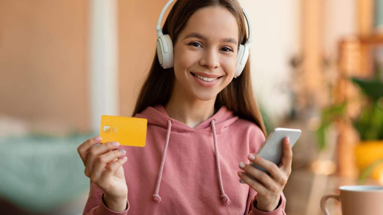 adolescente sorri com cartão de crédito e celular na mão e fones de ouvido