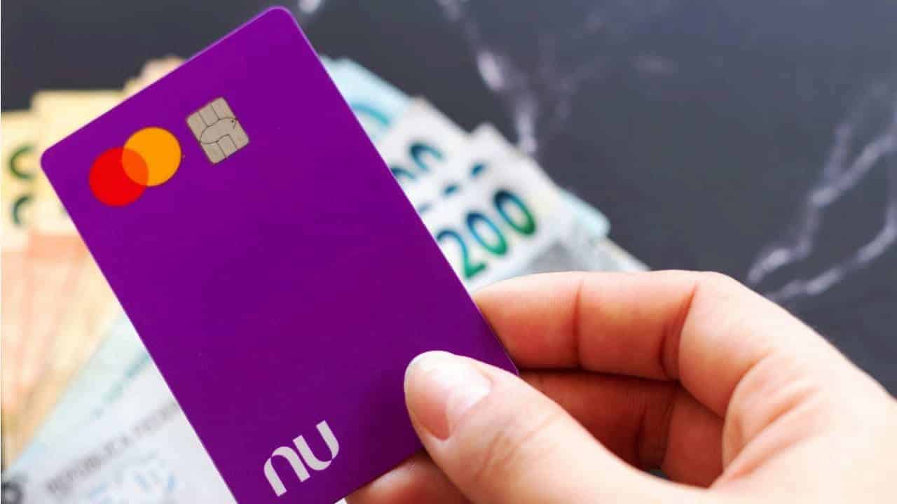 Pessoa segurando cartão do Nubank com notas de 200 reais ao fundo