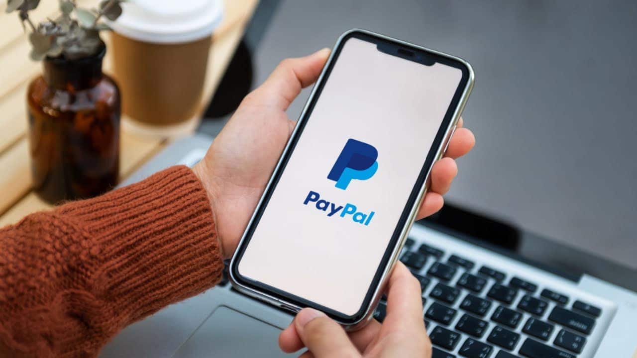 Mãos de uma pessoa segurando celular que mostra logo da carteira digital PayPal. Ao fundo, um notebook aberto.