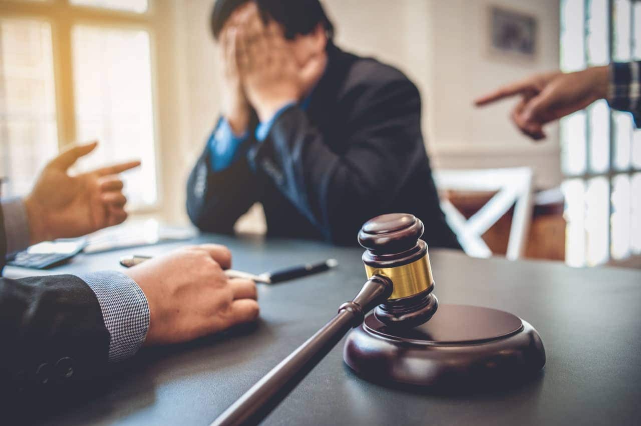 Homem com o rosto escondido nas mãos enquanto duas pessoas lhe apontam os dedos. Em primeiro plano, um malhete de juiz.
