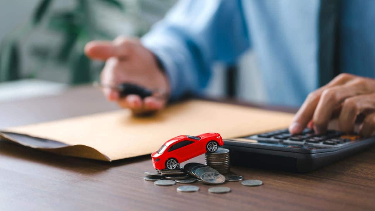 Em primeiro plano, miniatura de carro sobre montes de moedas ao lado de uma calculadora. Ao fundo, pessoa sentada segurando chave de carro.