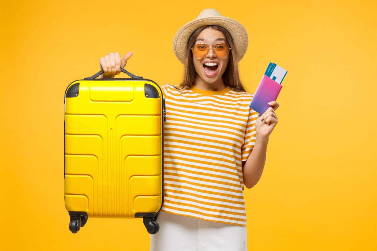 Mulher jovem sorrindo e segurando uma mala de viagem em uma mão e documentos na outra