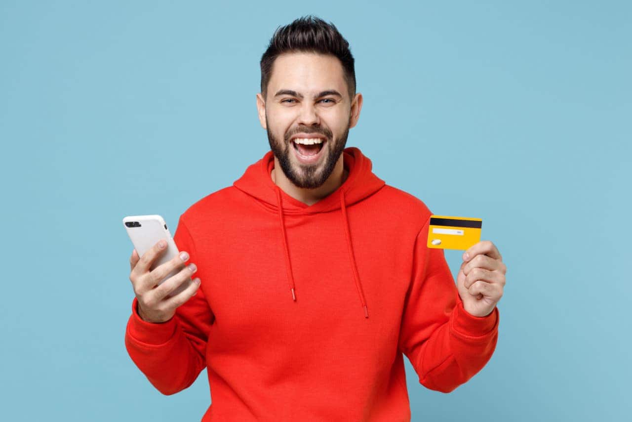 Homem sorridente comemorando com celular em uma mão e cartão de crédito na outra