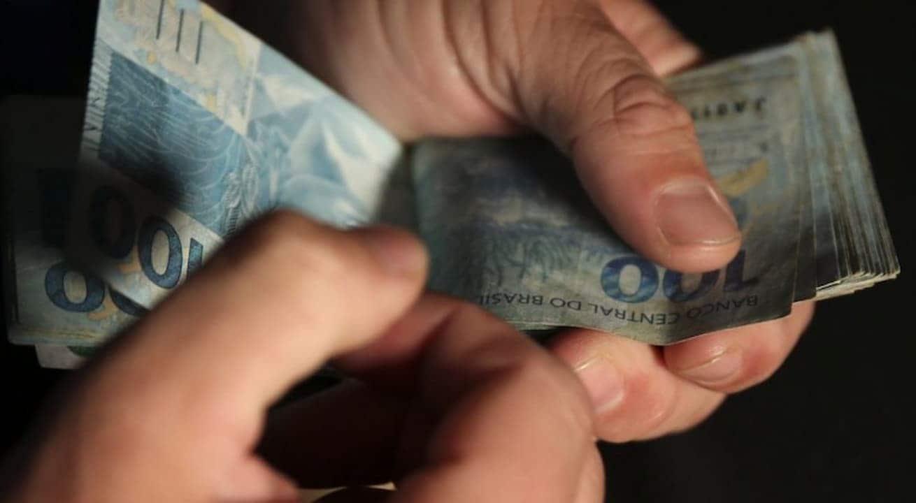 mãos contando notas de cem reais