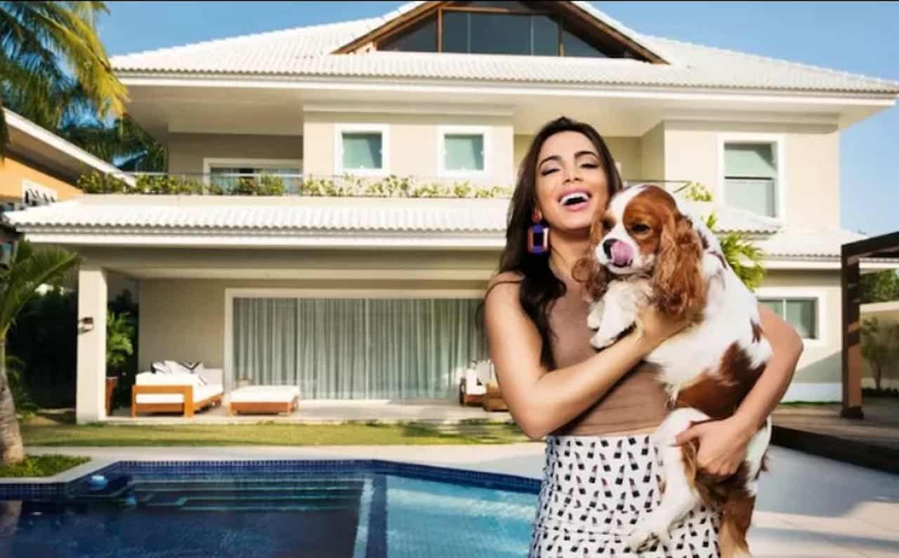 Segurando um cachorro, Anitta posa para um foto em frente a sua mansão