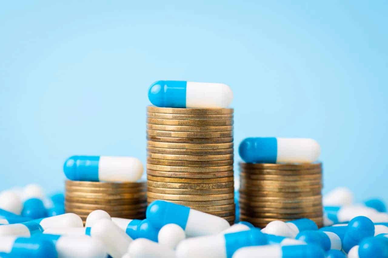 Três pilhas de moedas, cada uma com um cápsula de remédio azul e branca no topo, rodeadas por outras muitas cápsulas.