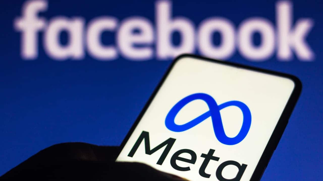 Sombra de uma mão segurando celular que mostra o logo da Meta. Ao fundo, o logo do Facebook.