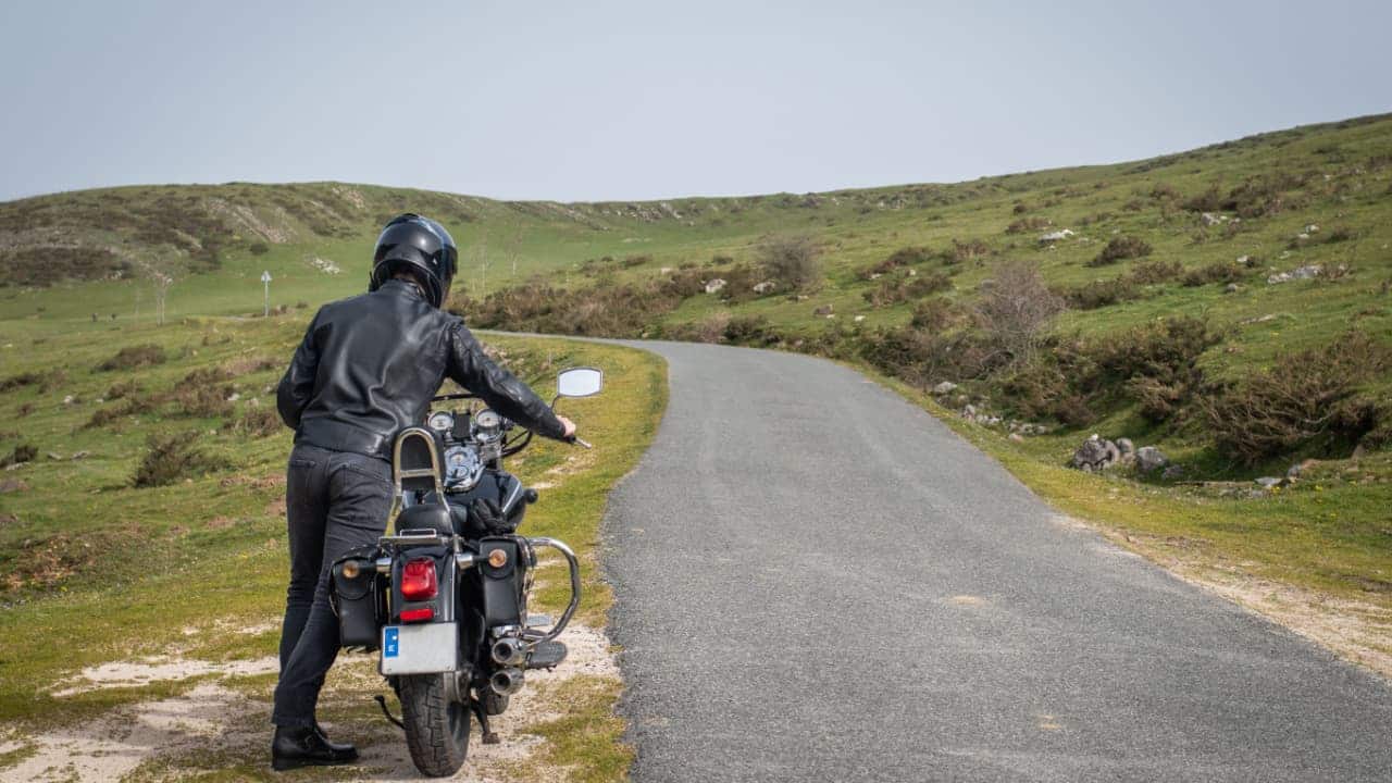 Jovem motociclista empurrando sua moto personalizada em uma estrada de montanha devido a uma falha no motor.