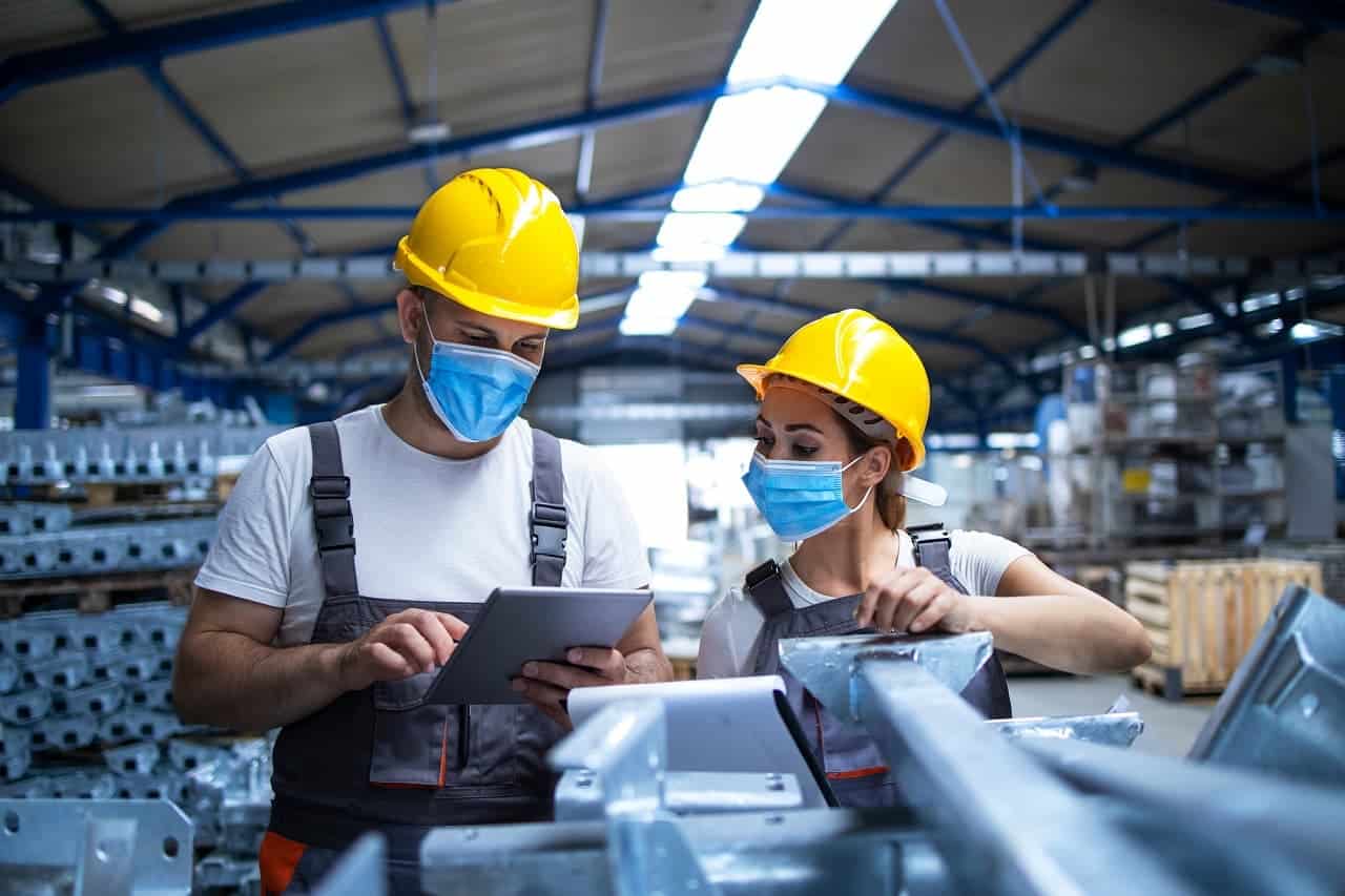 Trabalhadores industriais com máscaras faciais e equipamentos de segurança discutindo sobre a produção na fábrica.