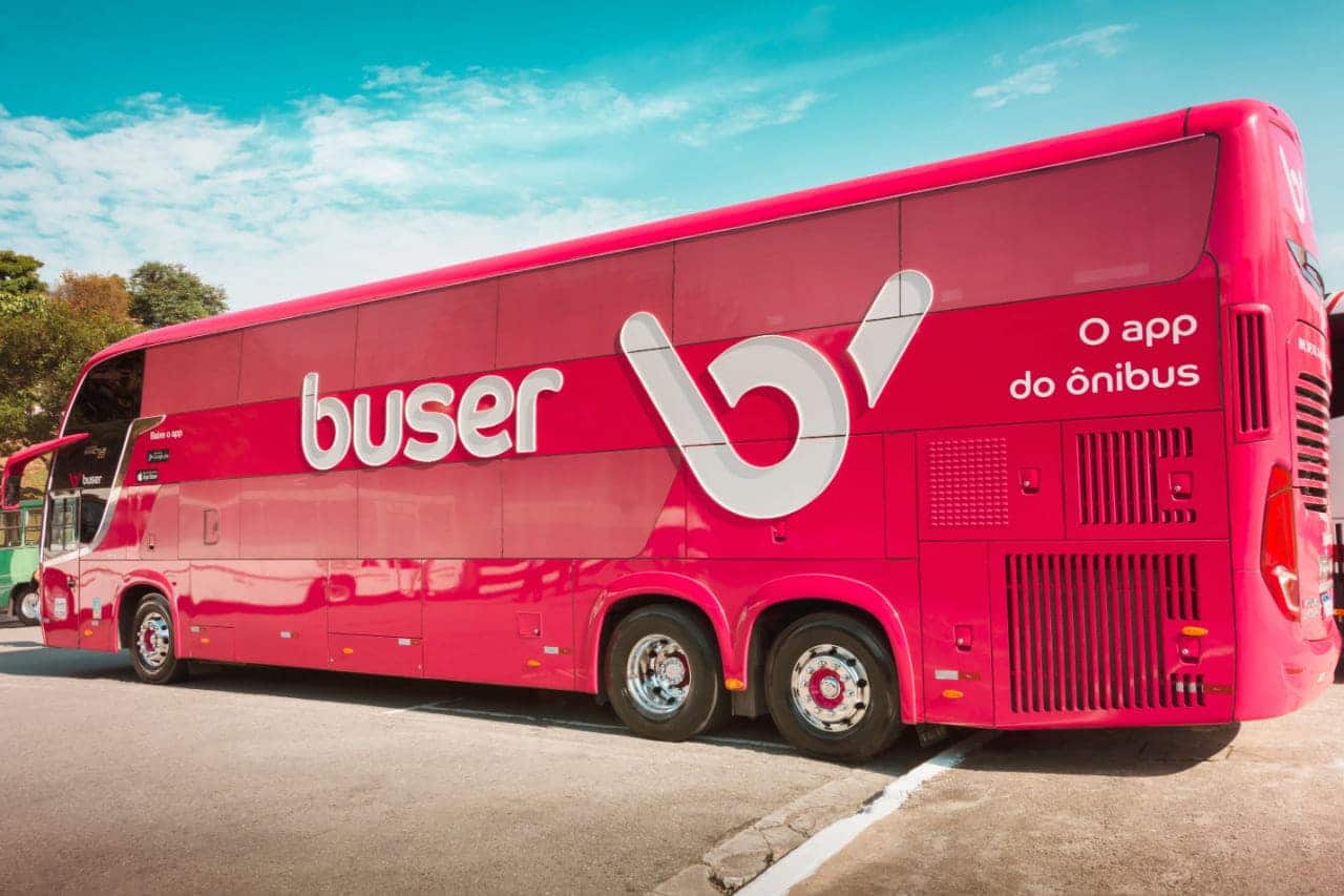 Lateral de um ônibus grande, da cor rosa, com logo e slogan da empresa Buser