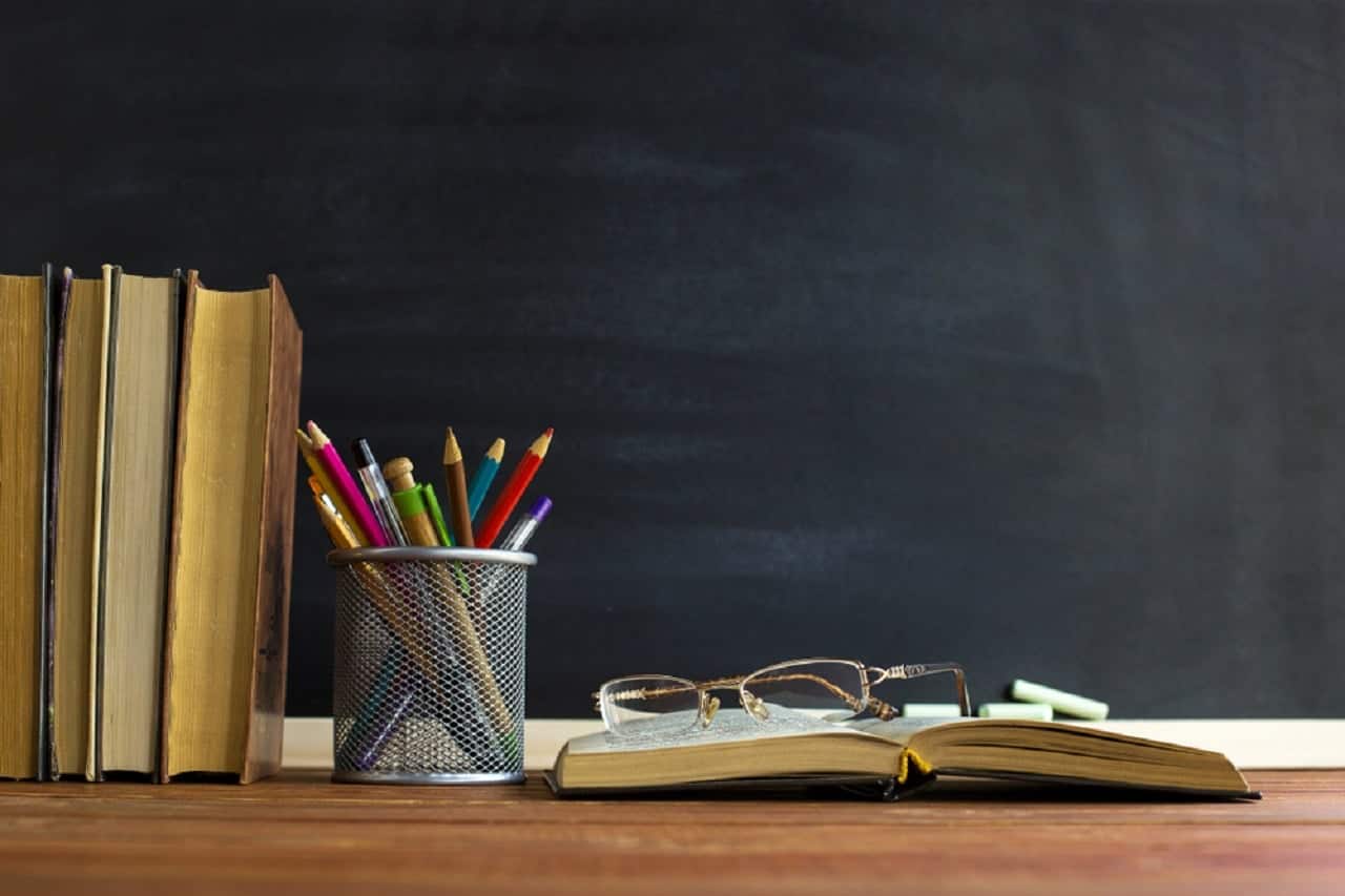 Imagem de uma mesa escolar com livro, lápis de cor e um óculos. Ao fundo, um quadro negro. edital pnd