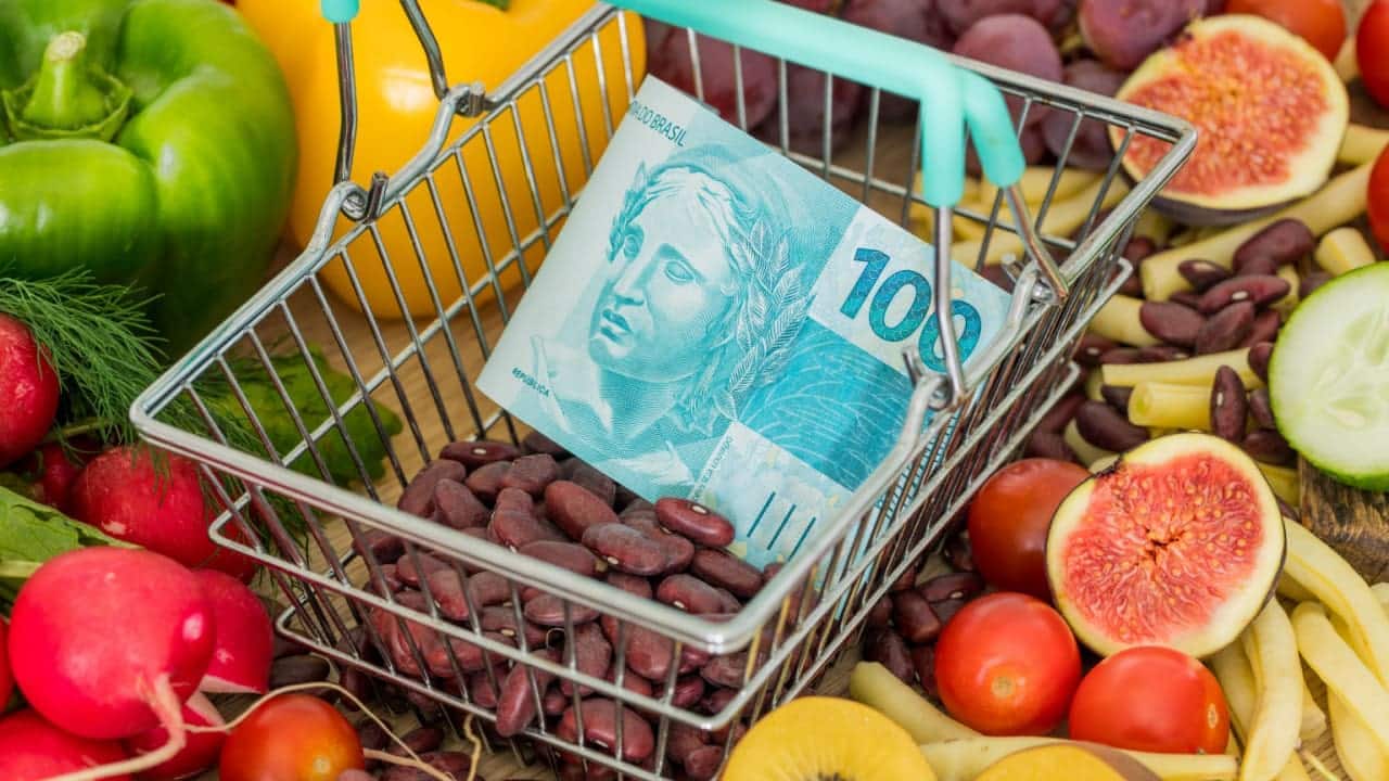 Nota de 100 reais dentro de uma cesta de compras rodeada de diversos tipos de alimentos, como frutas, verduras e grãos.