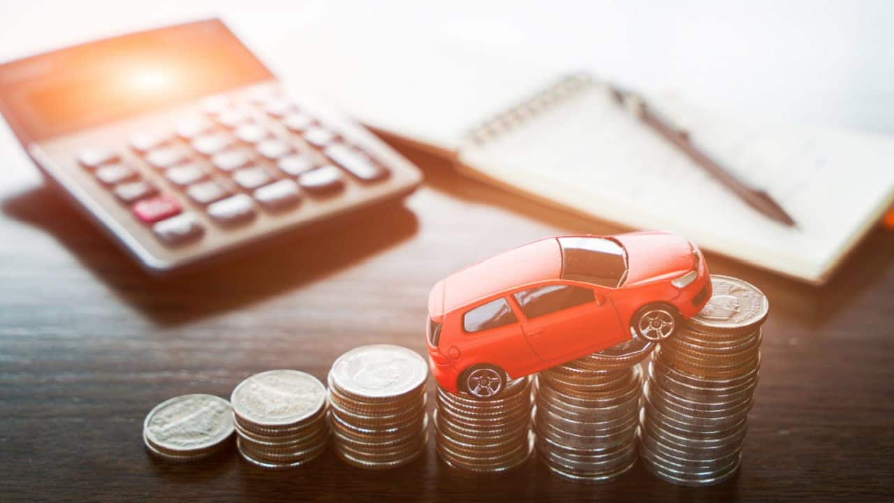 Sobre uma mesa, miniatura de um carro posicionada sobre pilhas de moedas organizadas em ordem crescente. Ao fundo, calculadora, caderneta e caneta.