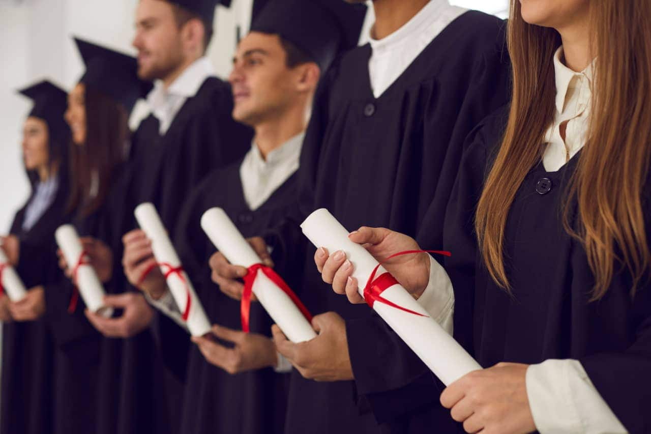 Estudantes de faculdade vestidos de beca, em fila, com diplomas nas mãos.