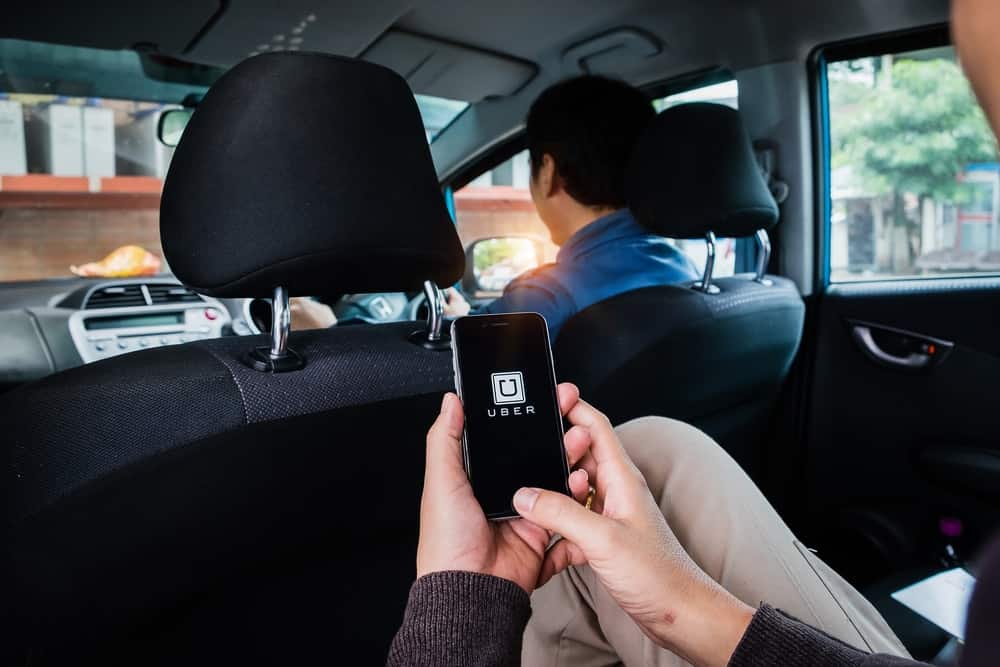 Imagem do interior de um automóvel. Um passageiro no banco traseiro está com o celular ligado mostrando o aplicativo da Uber.