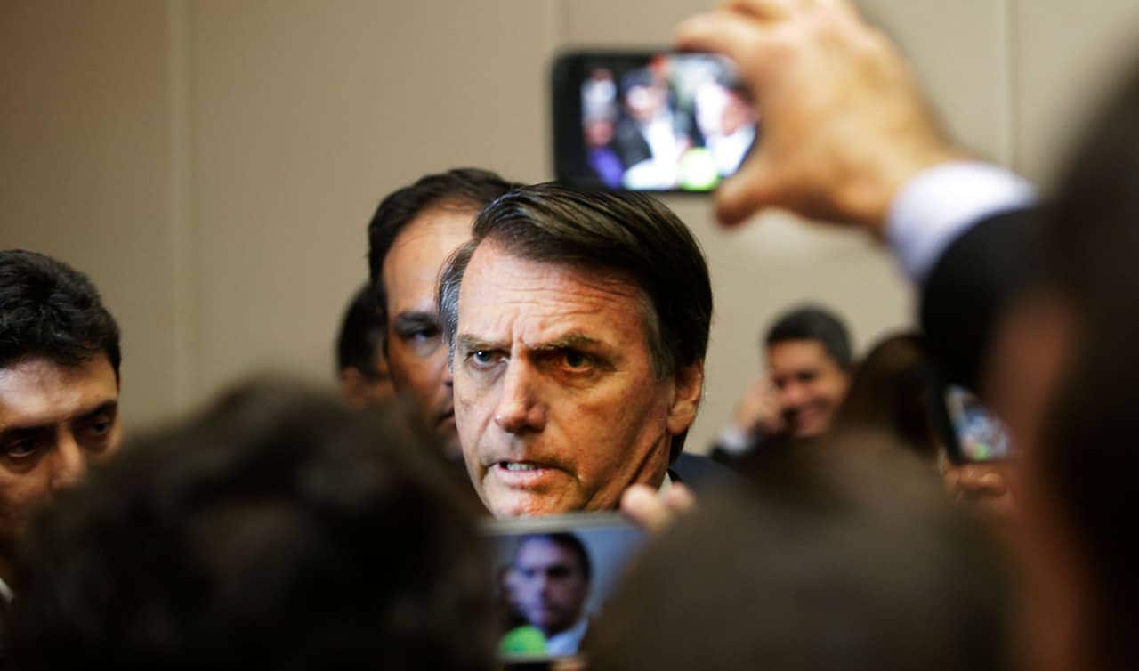 Jair Bolsonaro com expressão séria cercado por pessoas e câmeras de celular