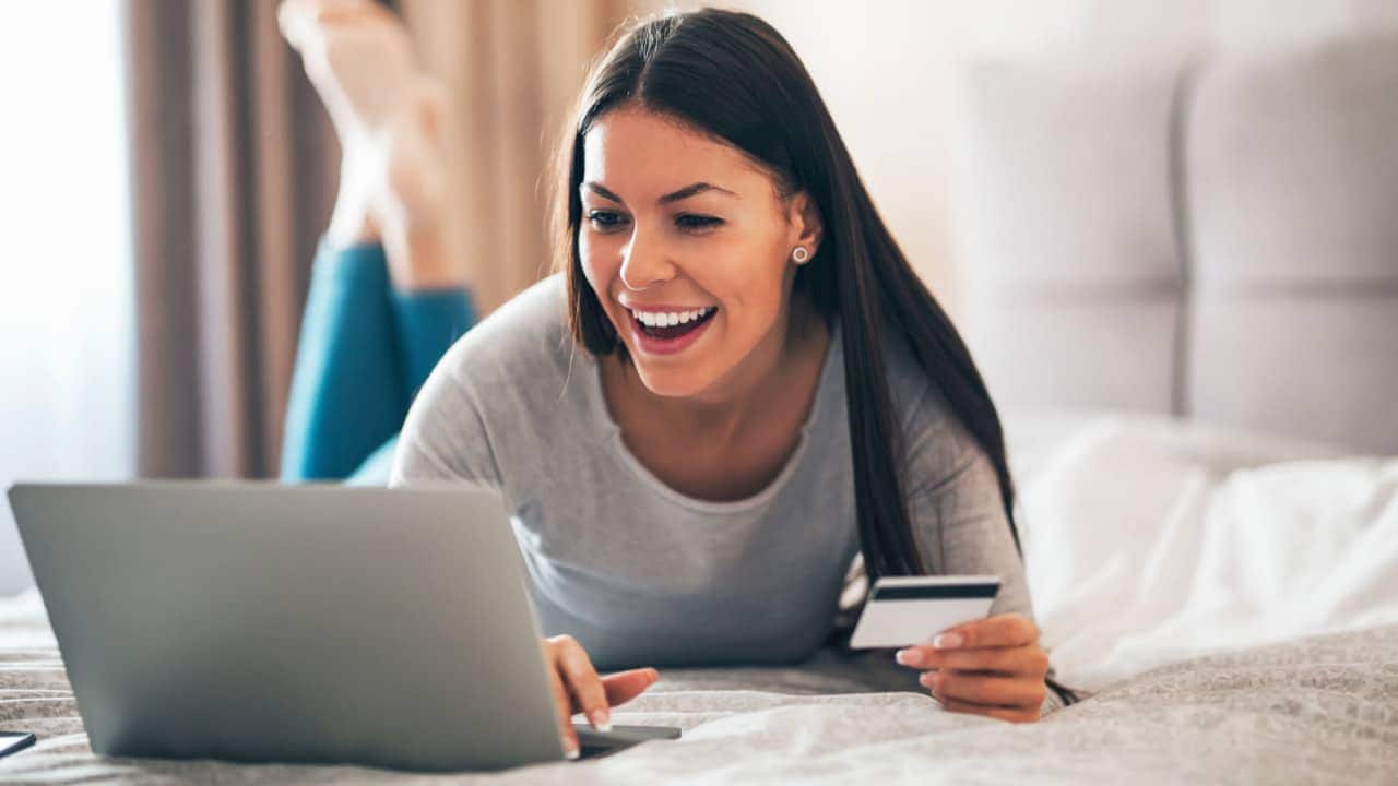 Mulher sorridente em uma cama, com as pernas para cima, enquanto utiliza um computador com uma das mãos. Na outra mão, segura um cartão de crédito.