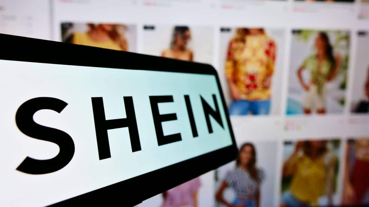 Pessoa segurando celular com o app da Shein aberto. Ao fundo, o catálogo de roupas femininas.