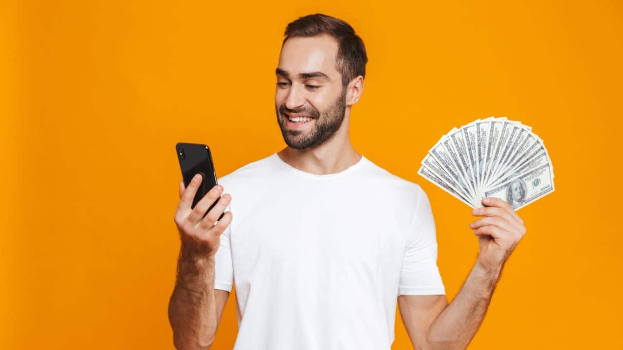Homem sorrindo e segurando celular com uma das mãos e notas de dinheiro com a outra