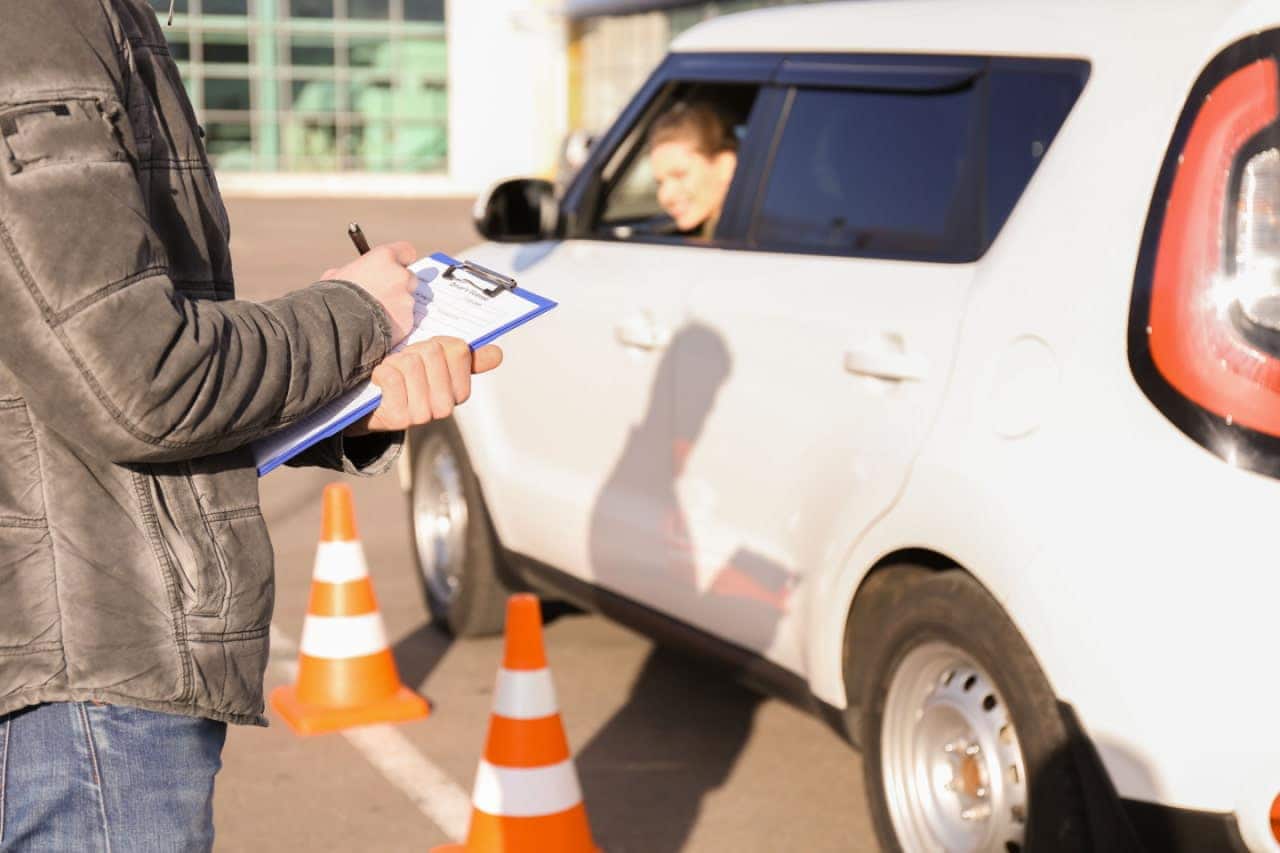 Condutora em veículo estacionado durante aula de autoescola. Ao lado do carro, dois cones. Em primeiro plano, instrutor fazendo anotações. Autoescolas