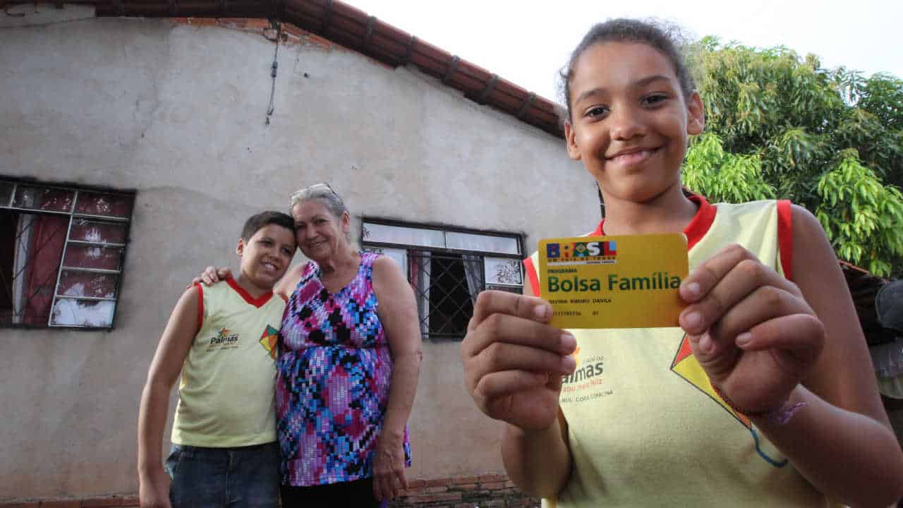 Menina segurando o cartão do bolsa família, ao fundo um menino e uma senhora em frente a uma casa