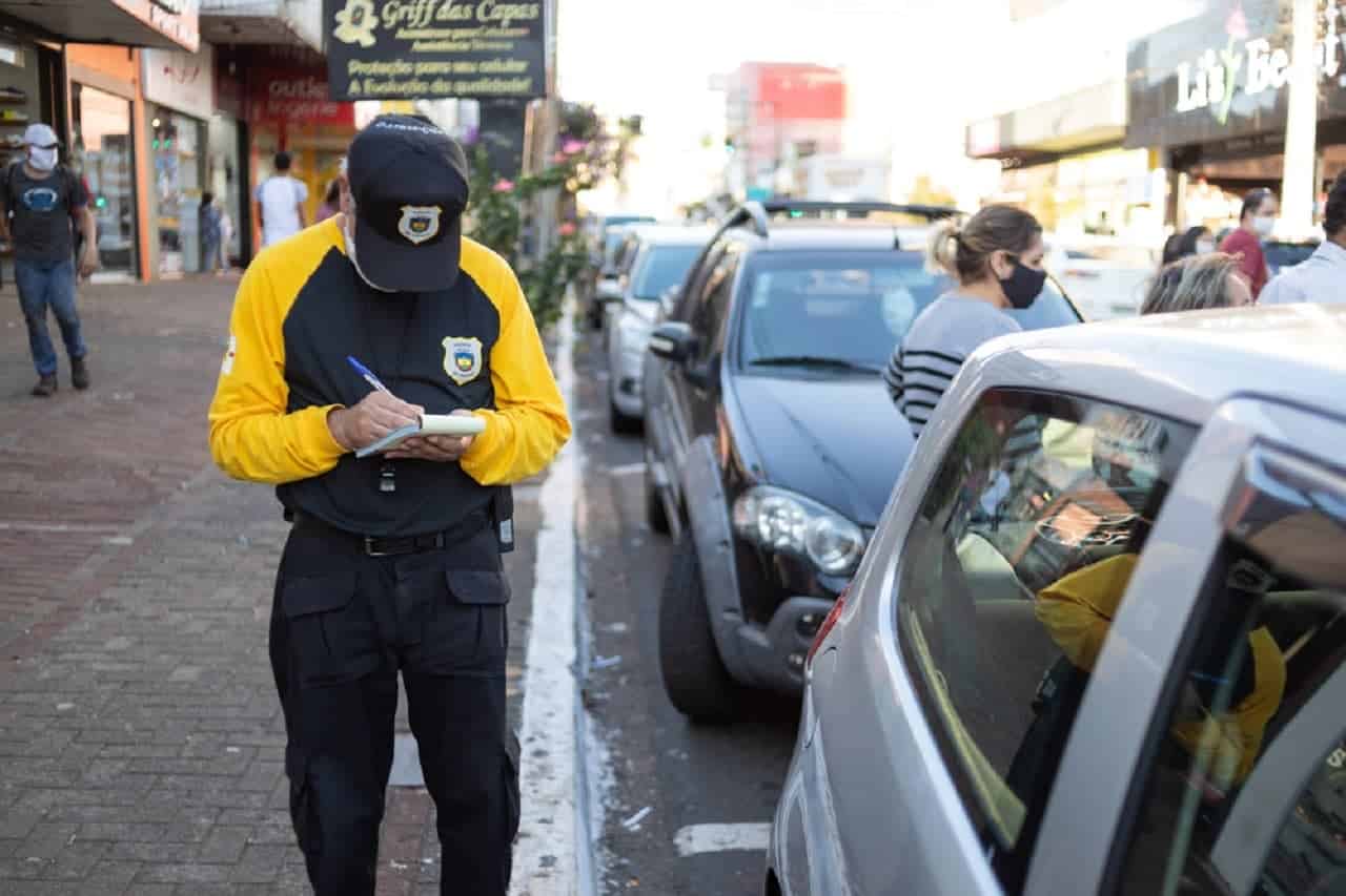 Imagem de um policial escrevendo uma multa. Ele está na calçada, há pedestres passando ao fundo e carros estacionados na via.