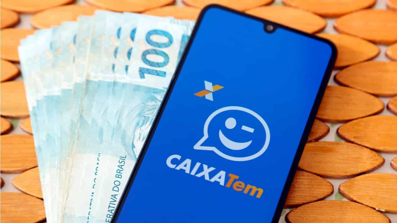 Celular exibindo aplicativo Caixa Tem ao lado de notas de 100 reais