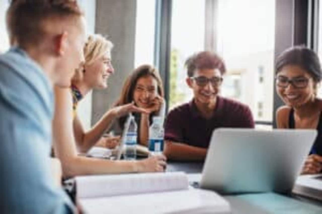 Quatro jovens reunidos em uma mesa rindo com computadores e cadernos