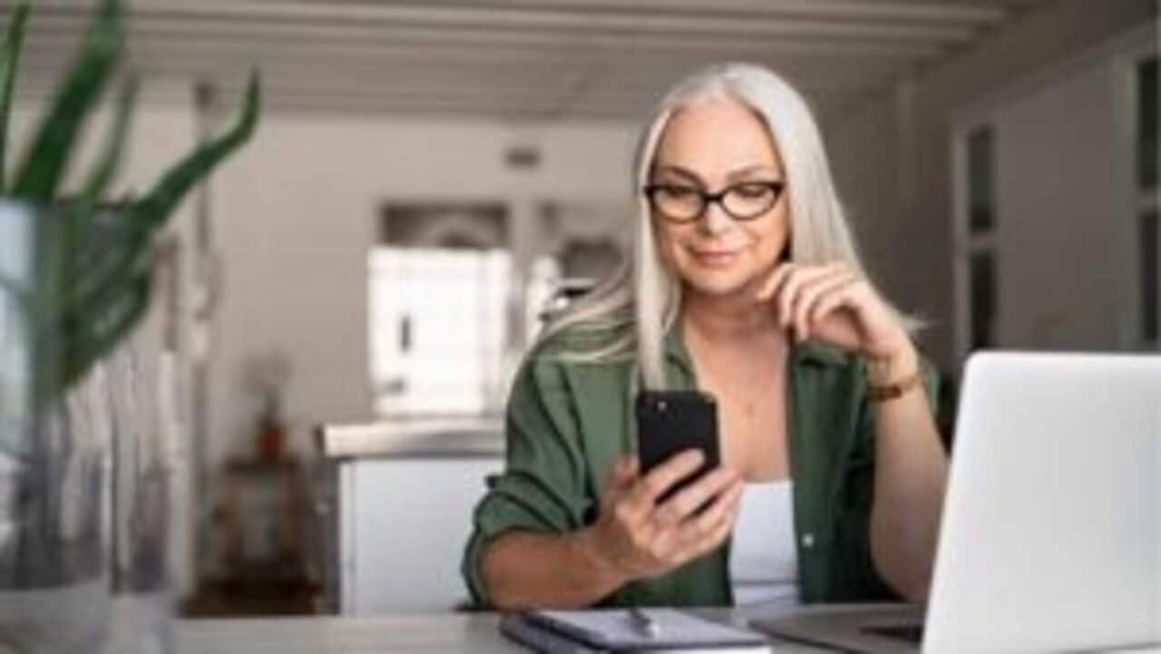 Mulher de cabelos longos e brancos utilizando Ăłculos e camiseta verde sentada em uma mesa segundando o celular