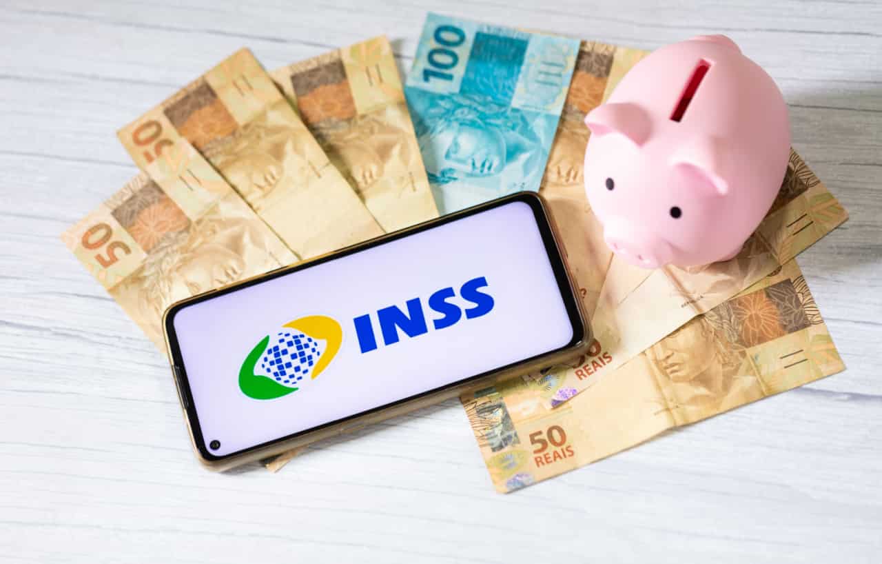 Smartphone próximo a notas de real e um cofre em forma de porquinho exibe o logo do INSS