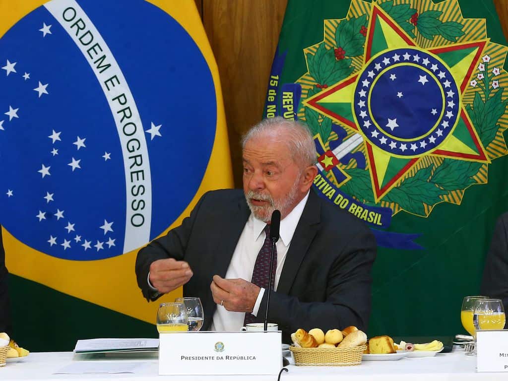 Lula em frente a bandeira do Brasil discursando.