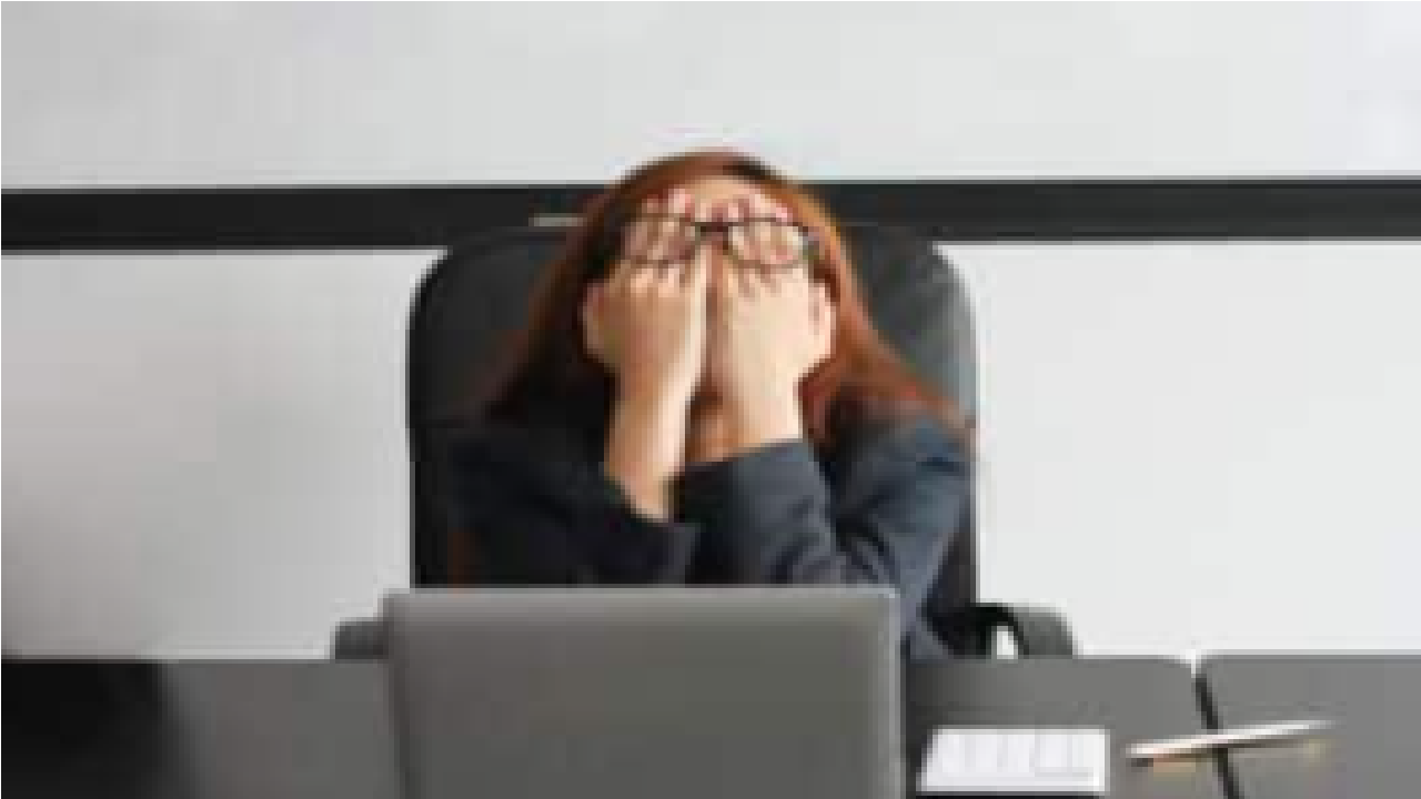 Imagem de mulher sentada em mesa de escritório com as mãos embaixo dos óculos apoiadas nos olhos