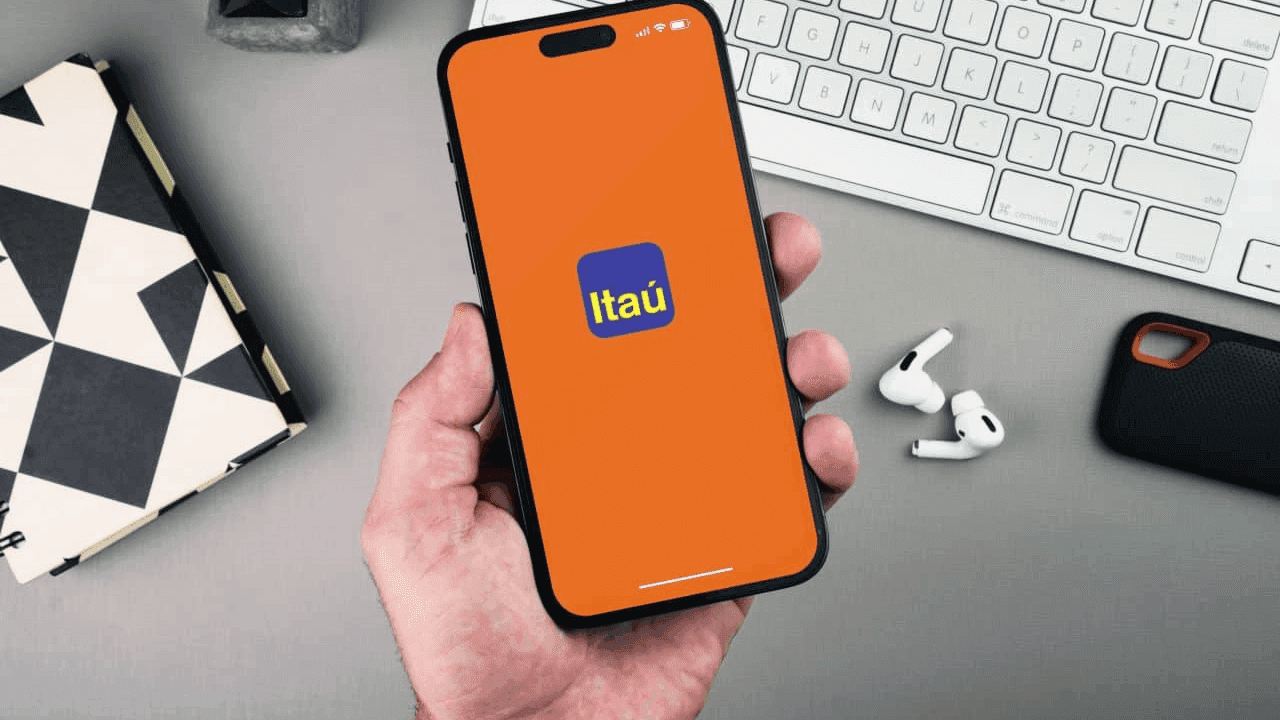 Imagem de mão masculina segurando celular que mostra na tela logo do Banco Itaú em fundo laranja
