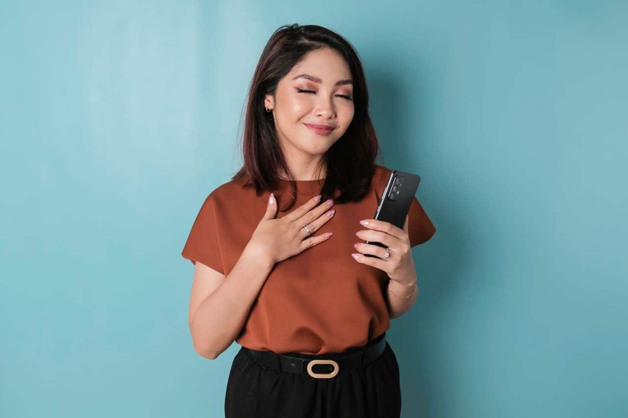 Mulher segurando celular sorrindo de olhos fechados com uma mão no coração