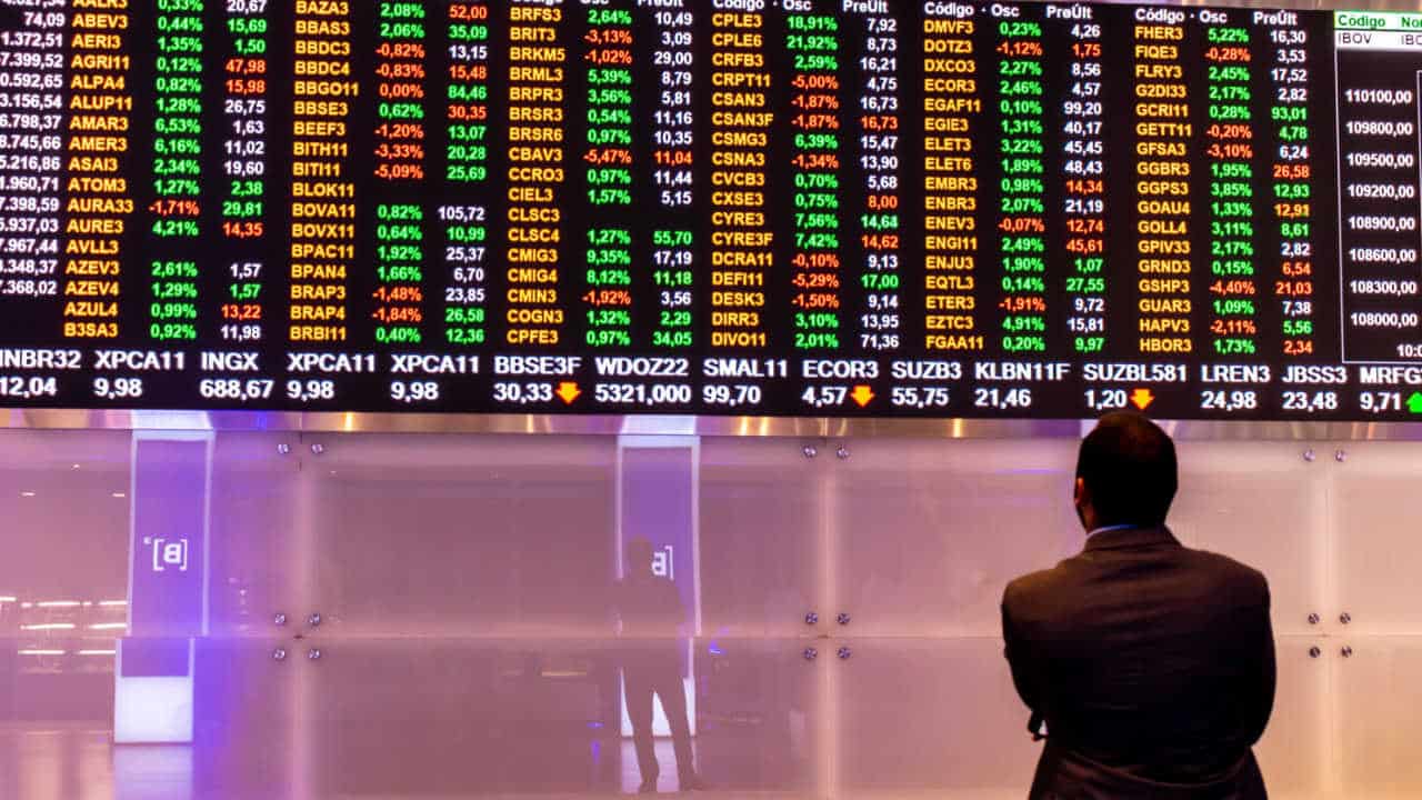 Homem de pé observando tela que mostra cotação de ações da Bolsa de Valores fechamento