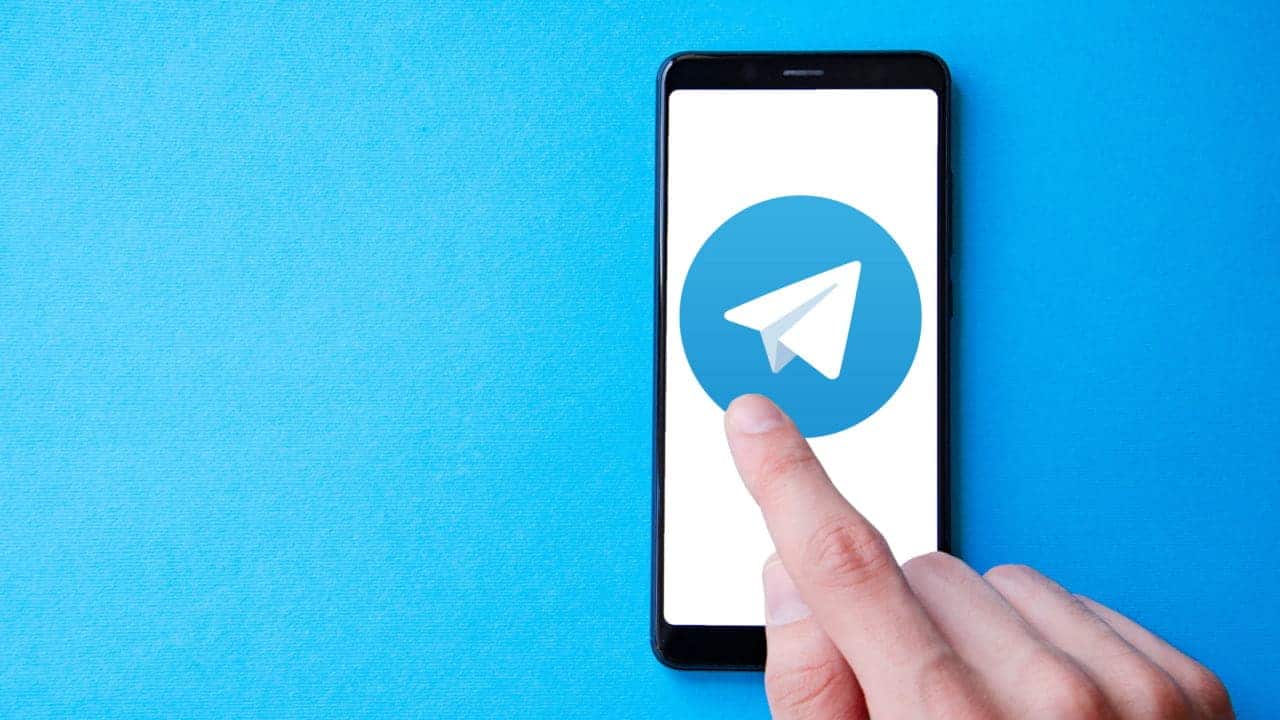 Celular que mostra logo do aplicativo de mensagens Telegram sobre uma superfície azul. No canto inferior direito, uma mão toca a tela com o dedo indicador.