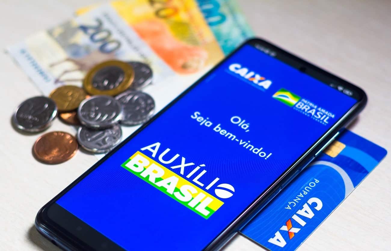 Celular com o aplicativo do Auxílio Brasil aberto. Debaixo do celular há um cartão da Caixa e ao lado esquerdo notas e moedas de Real