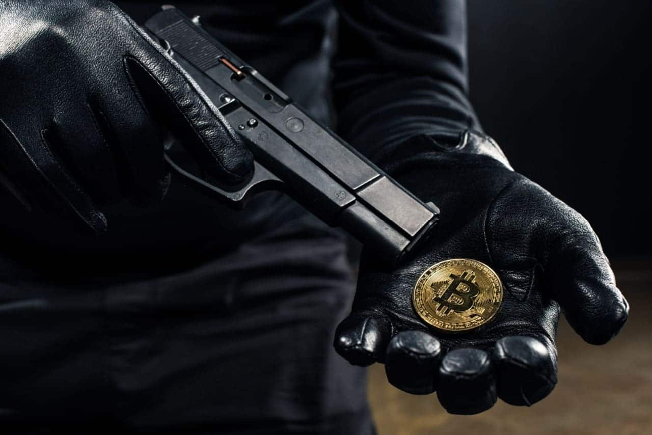 Pessoa segurando moeda de bitcoin em uma das mãos e apontando uma arma para o objeto com a outra mão. A pessoa está de luvas e roupas pretas.